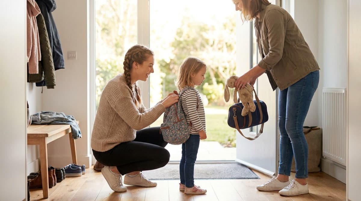 Ein Au-Pair hockt vor einem Mädchen und richtet den Rucksack, während die Mutter an der Haustür eine Tasche reicht